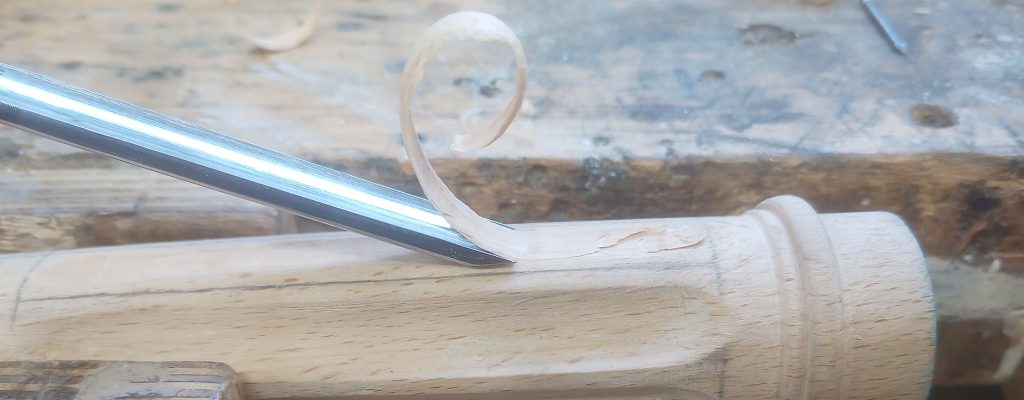A strip of wood curls up from a turned chair leg where it's being removed with a chisel.