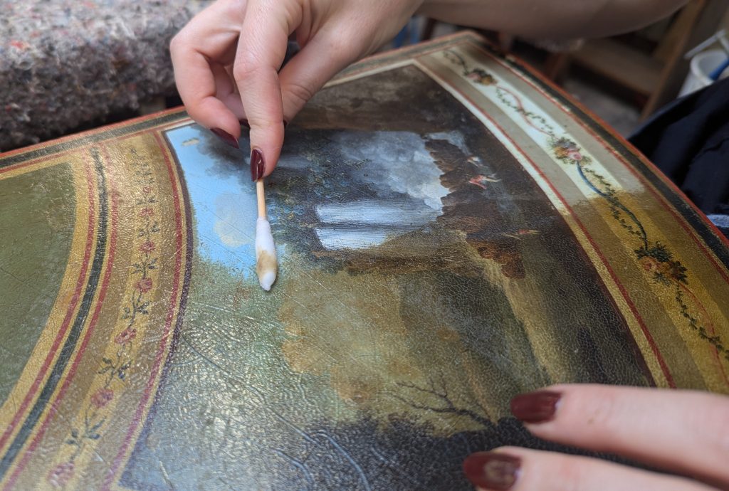 A woman with varnished nails removes discoloured lacquer from a painted antique table with a cotton bud.