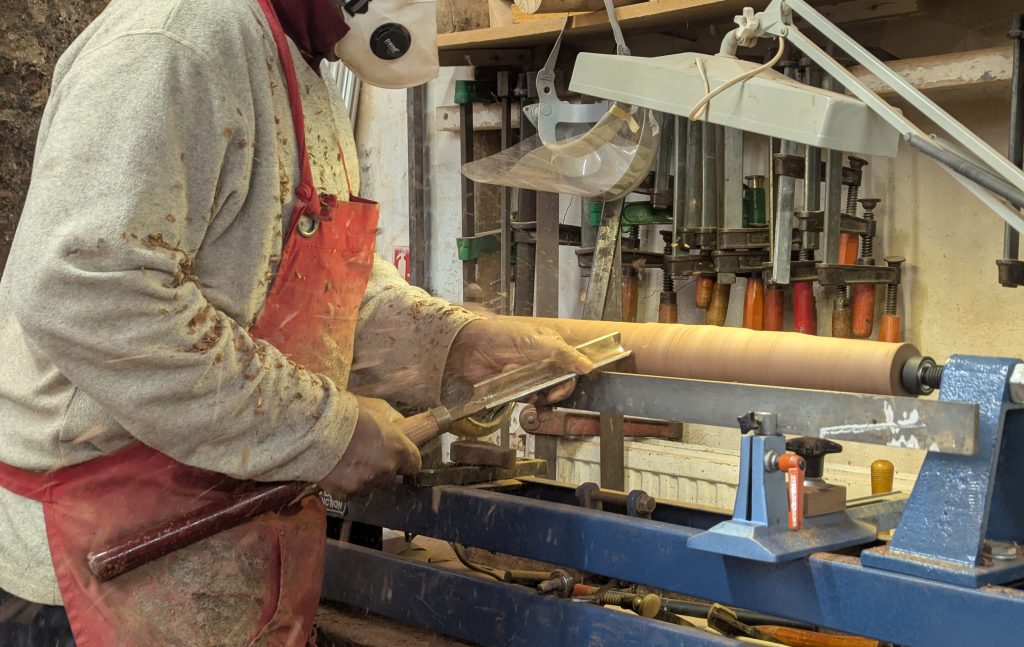 A man in an orange apron carves a wooden table leg on a lathe with a chisel.